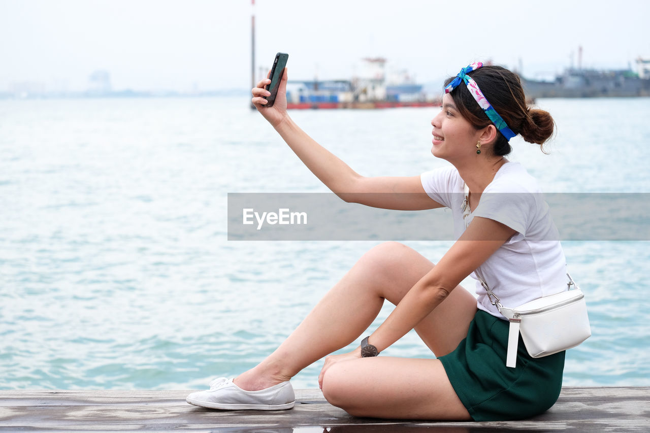 Side view of woman taking selfie while sitting at harbor