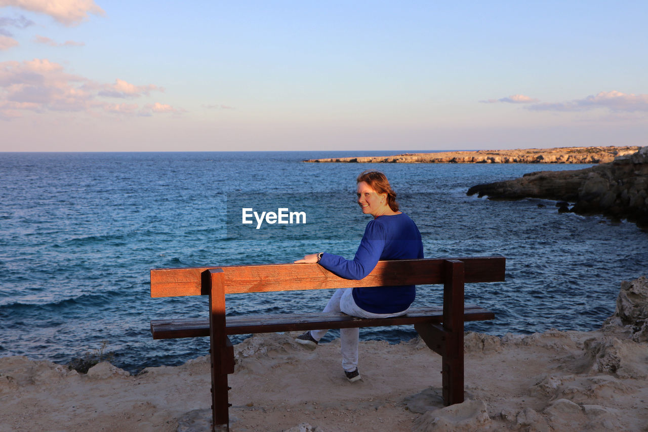 Portrait of woman sitting on bench looking at sea shore against sky