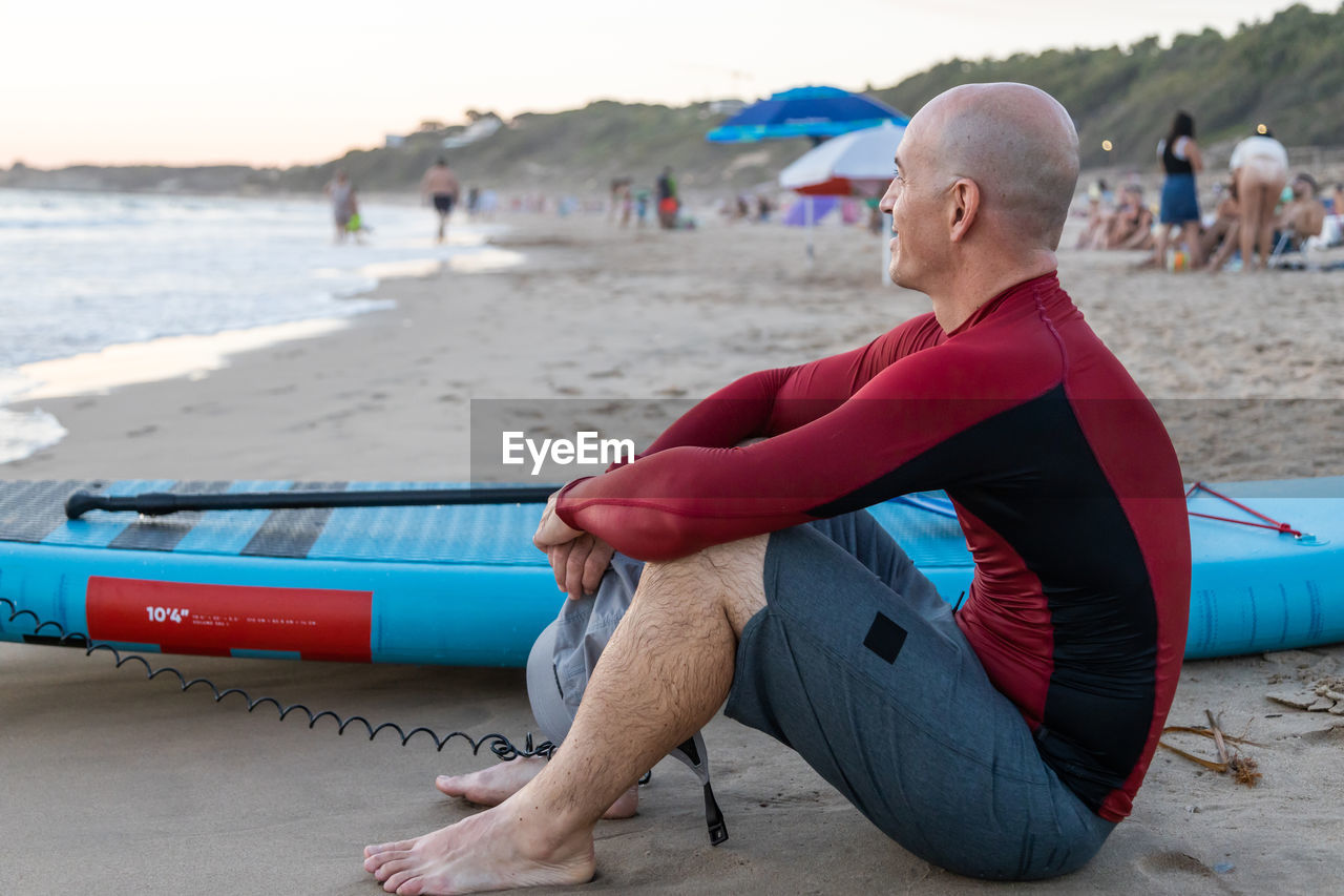 Side view of thoughtful male surfer in wetsuit sitting looking away with sup board while preparing to surf on seashore