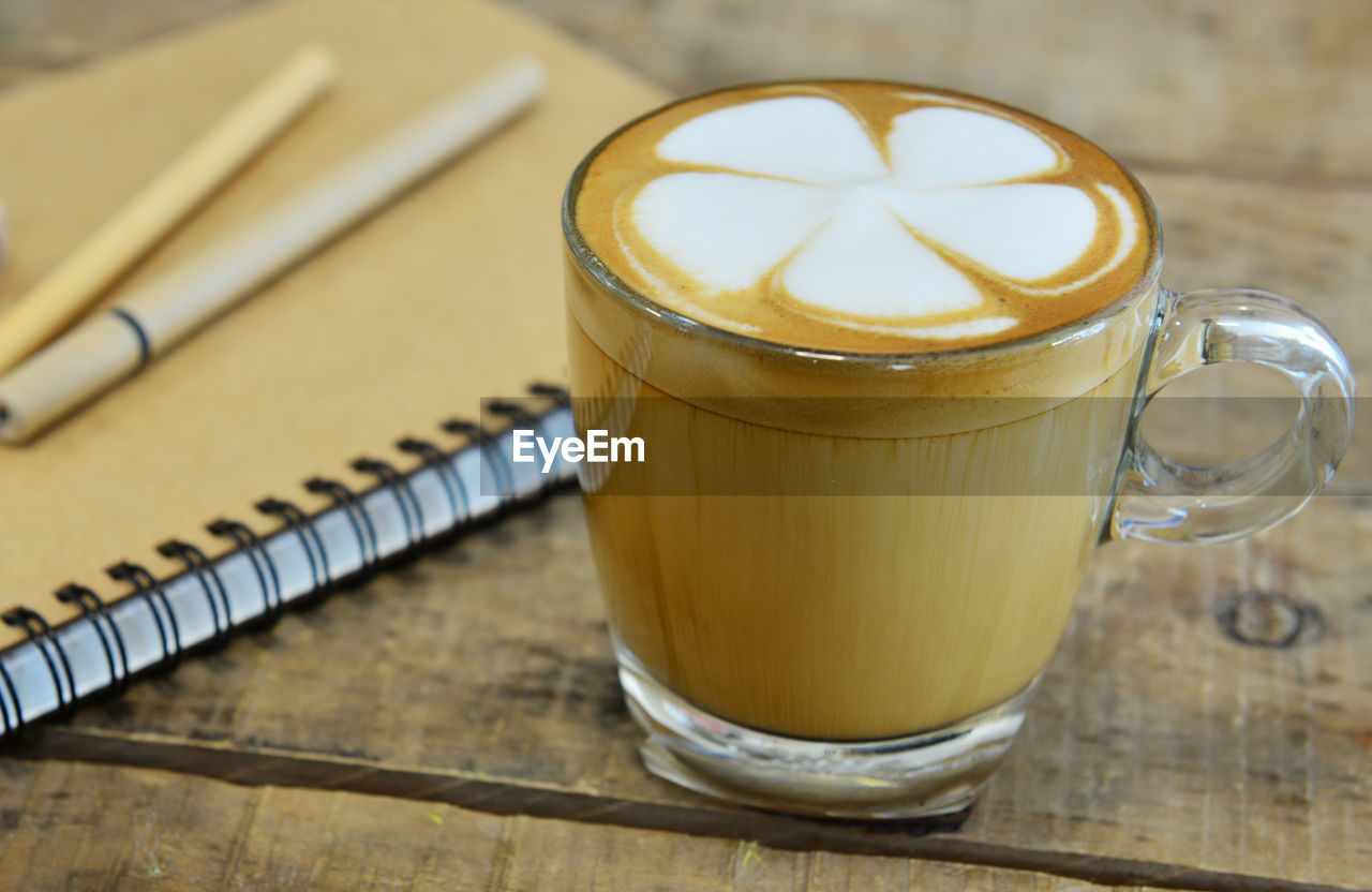 Close-up of frothy coffee by book and pen on wooden table