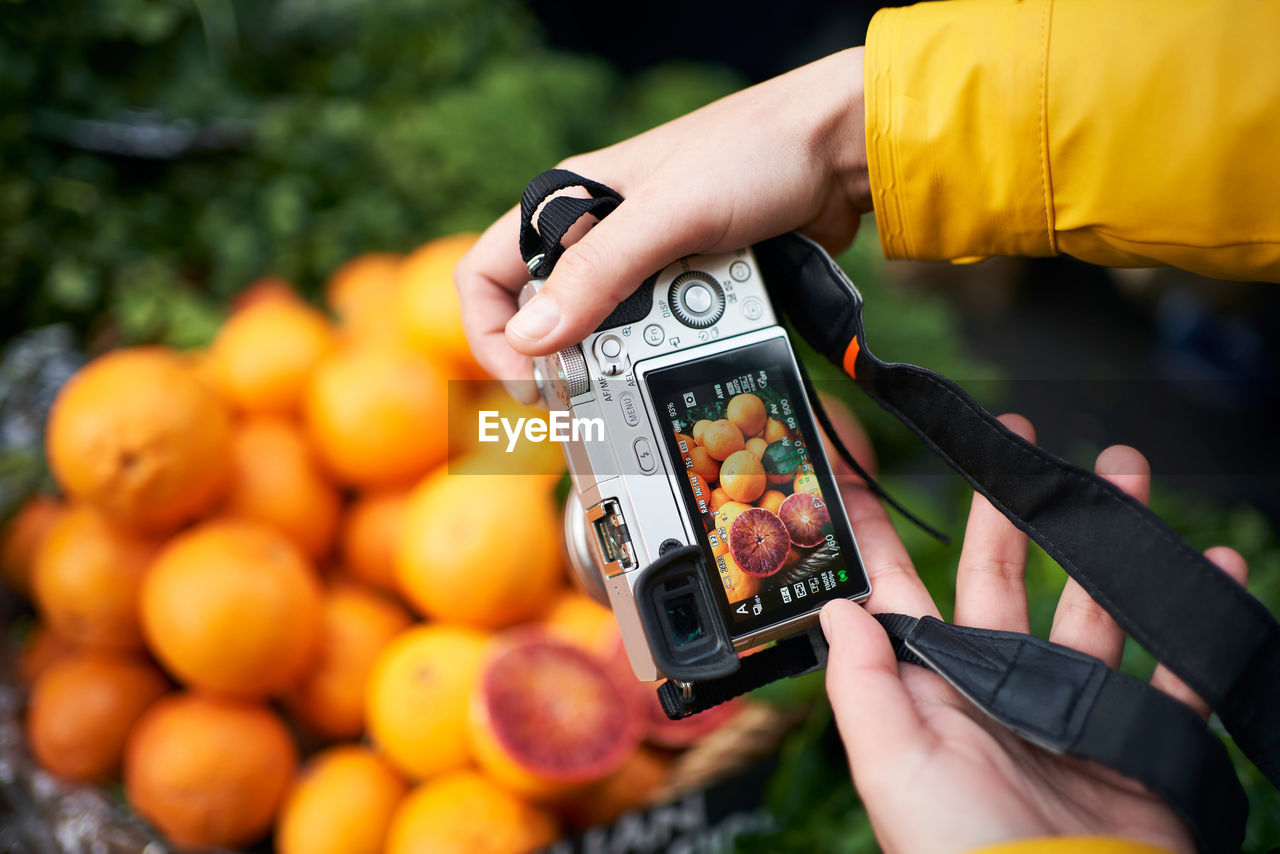 From above of crop anonymous person with digital photo camera taking picture of ripe orange fruits while visiting grocery market