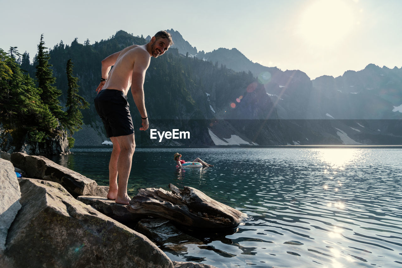 Portrait of shirtless mature man standing on rock at lakeshore against sky during sunny day