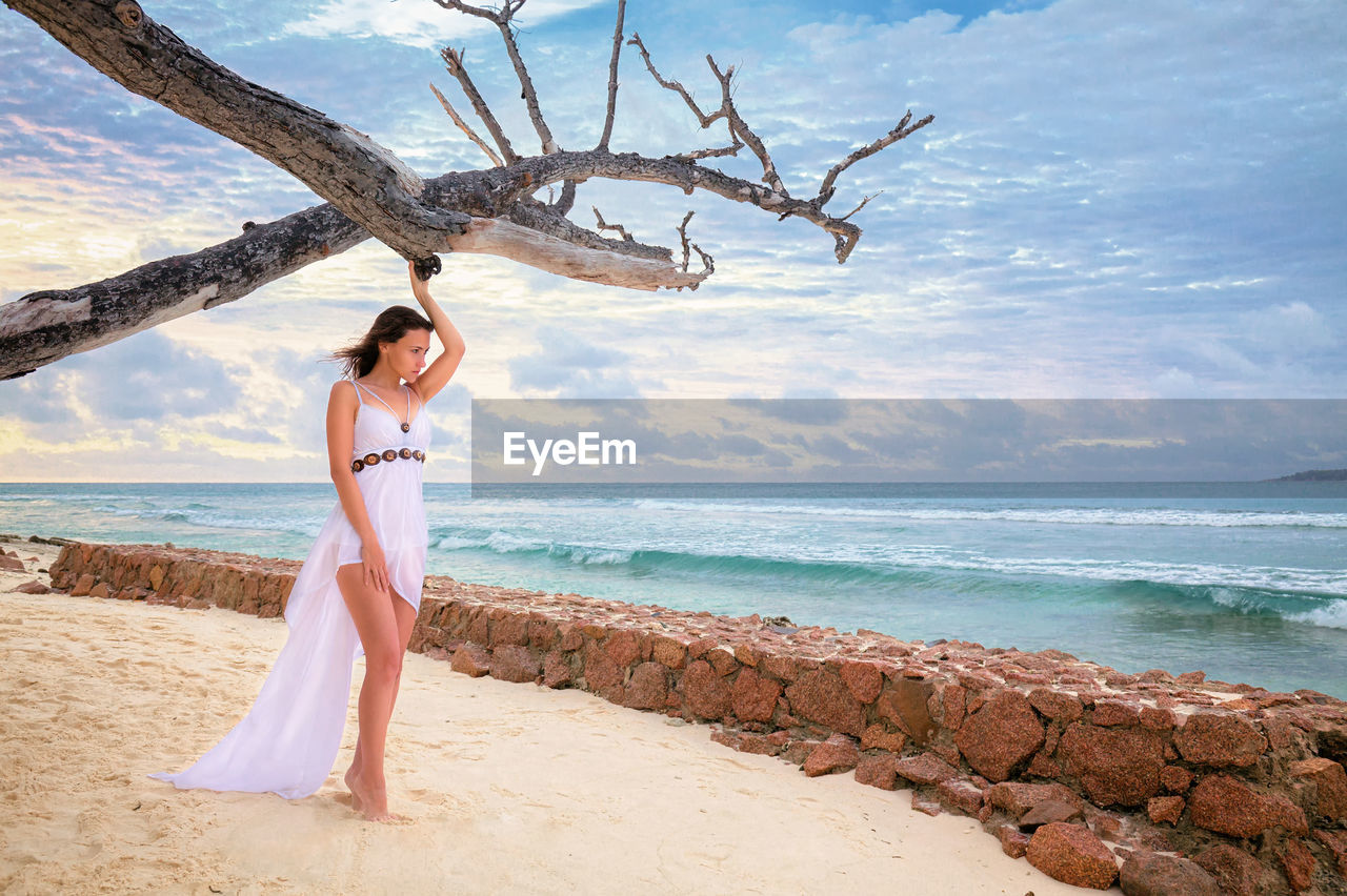 Full length of woman wearing white dress standing at beach against sky