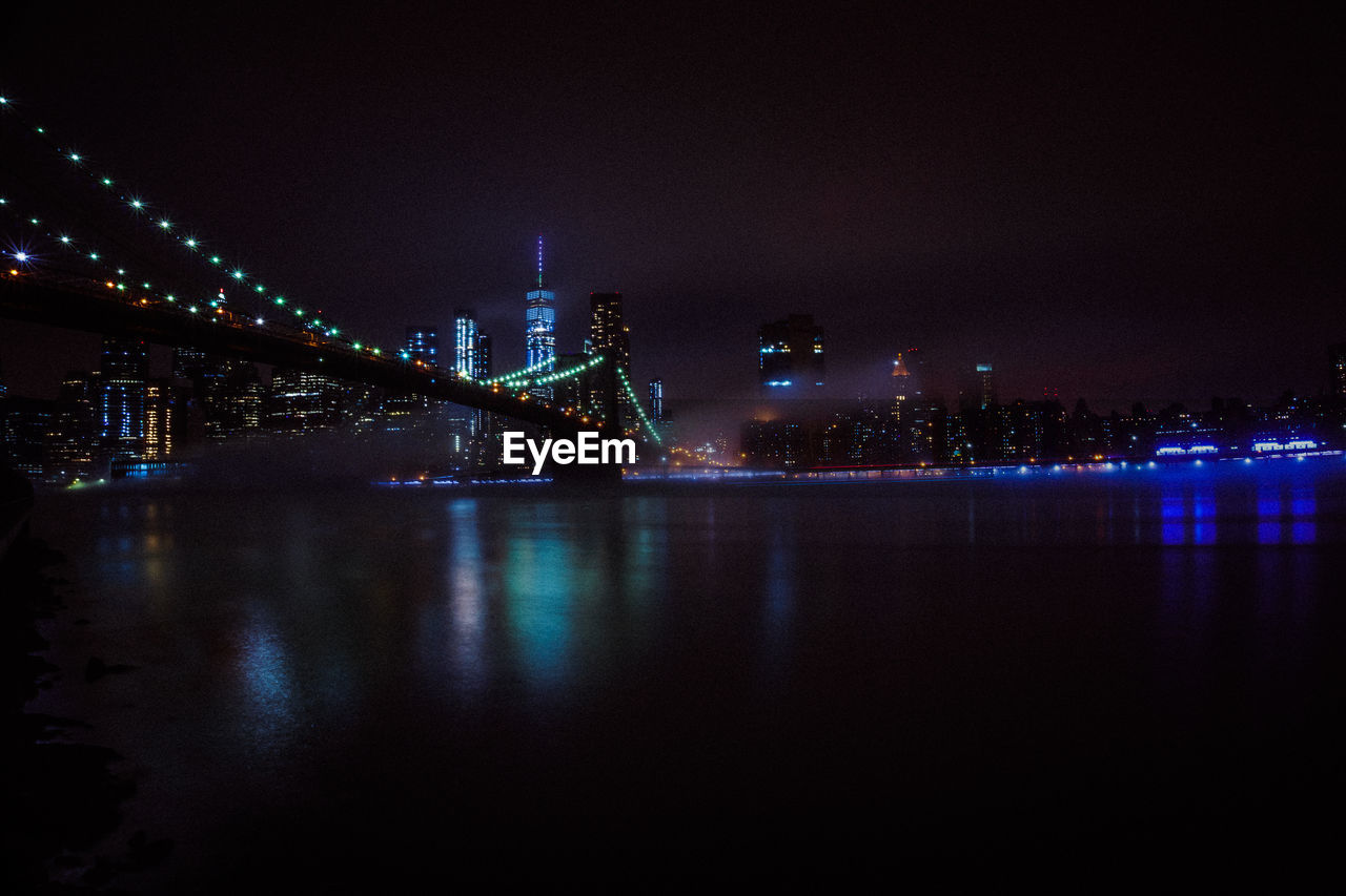 Illuminated modern buildings by river against sky at night