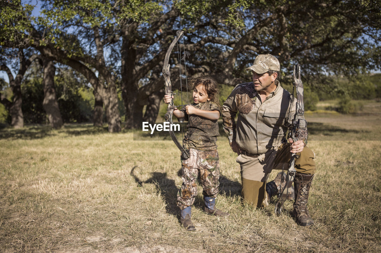 Girl aiming with arrow while father guiding on grassy field