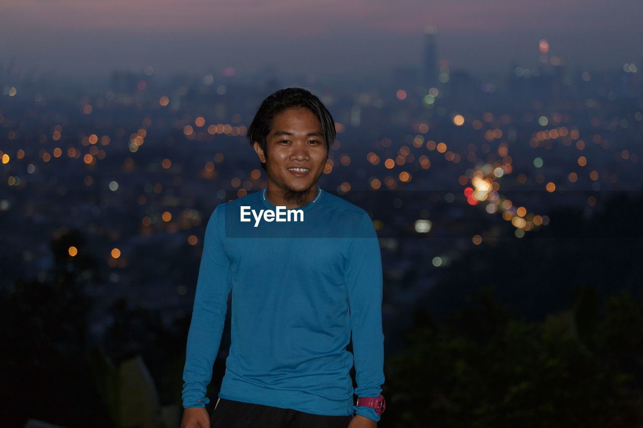 Portrait of smiling man standing against illuminated city at dusk