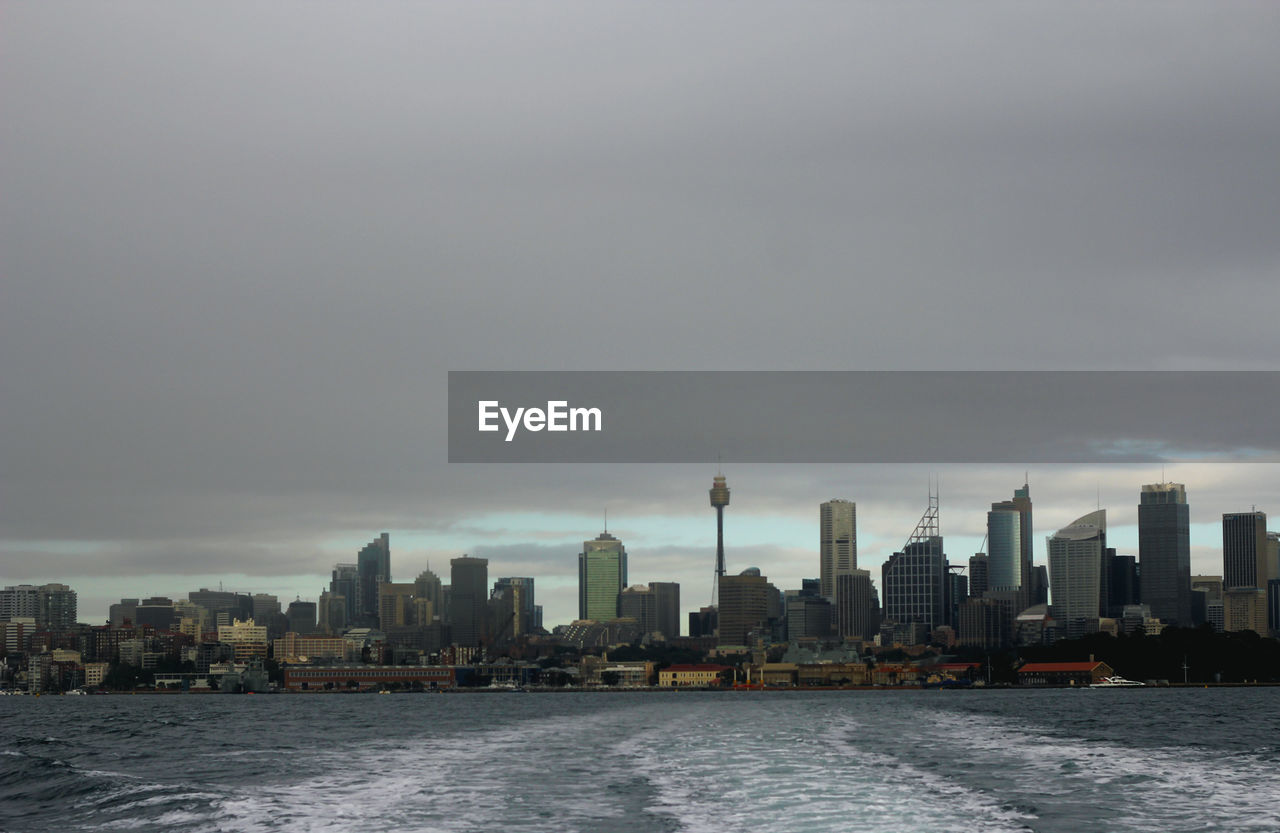 Cityscape by sea against cloudy sky