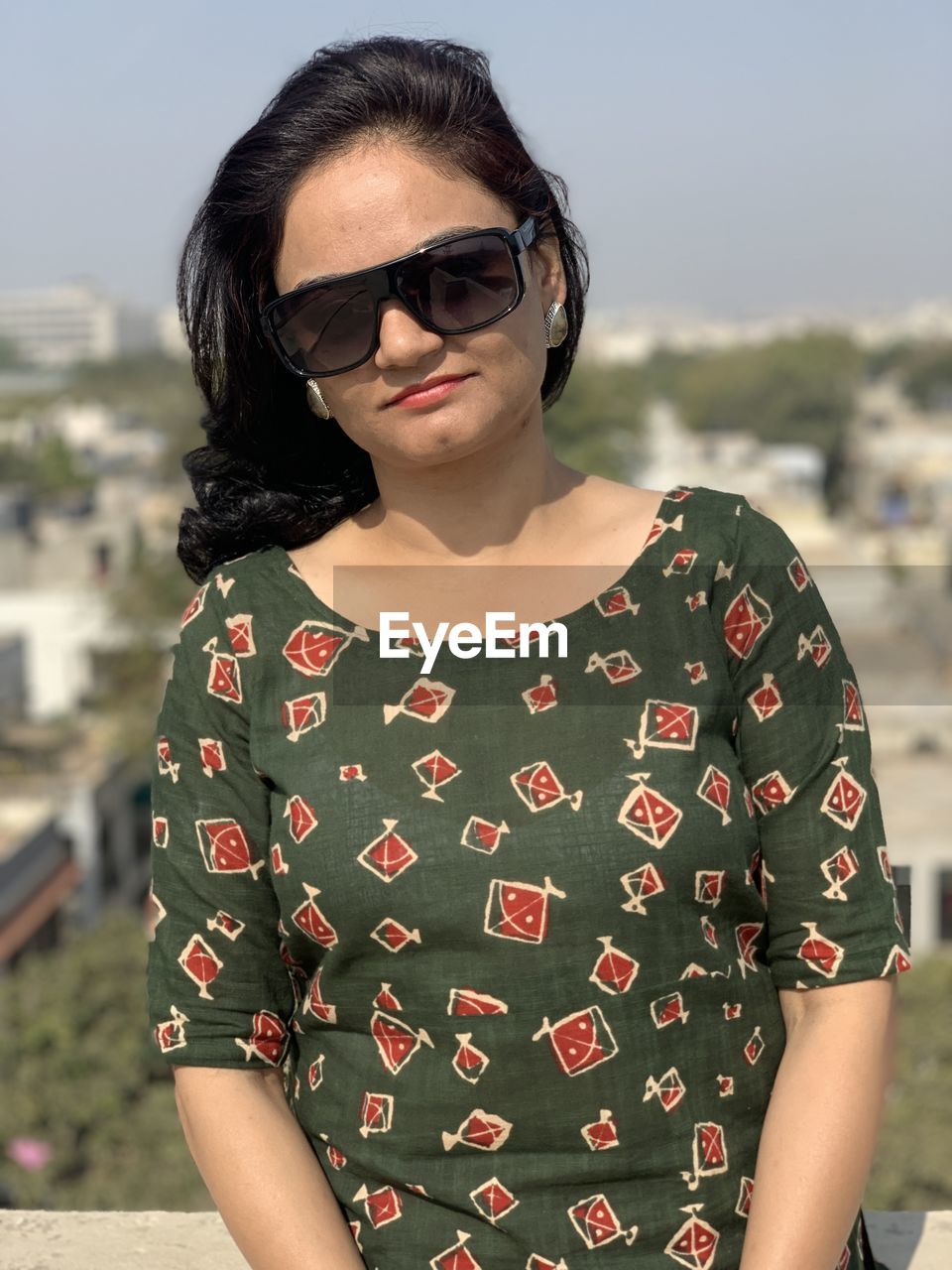 fashion, sunglasses, glasses, one person, front view, portrait, pattern, adult, spring, standing, young adult, women, waist up, photo shoot, hairstyle, dress, focus on foreground, nature, casual clothing, clothing, day, sleeve, black hair, brown hair, lifestyles, looking at camera, outdoors, architecture, cool attitude, leisure activity, person, sunlight, polka dot, sky, emotion, smiling, long hair, arts culture and entertainment, human face, city, female, three quarter length, sunny