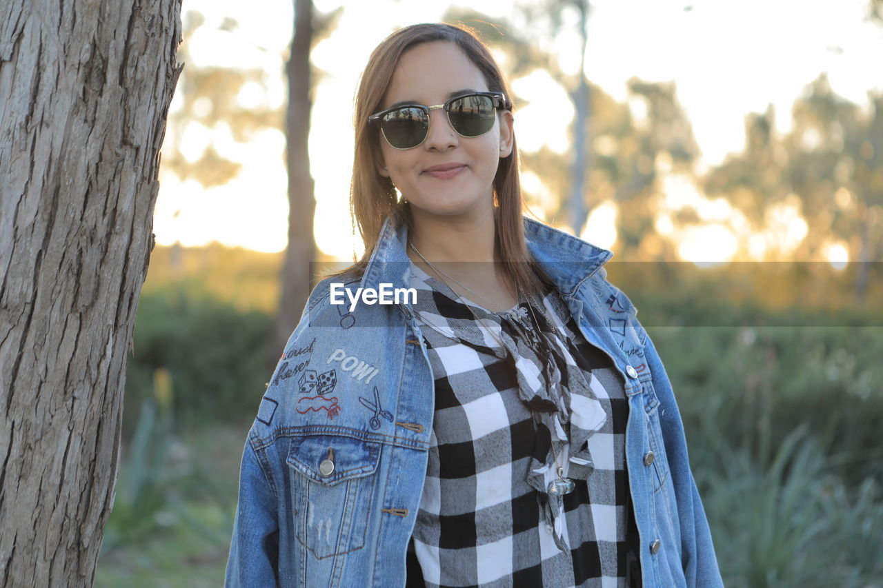 PORTRAIT OF SMILING WOMAN STANDING BY TREE TRUNK