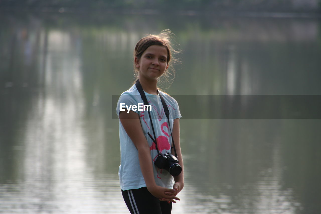 Portrait of girl with camera standing against lake