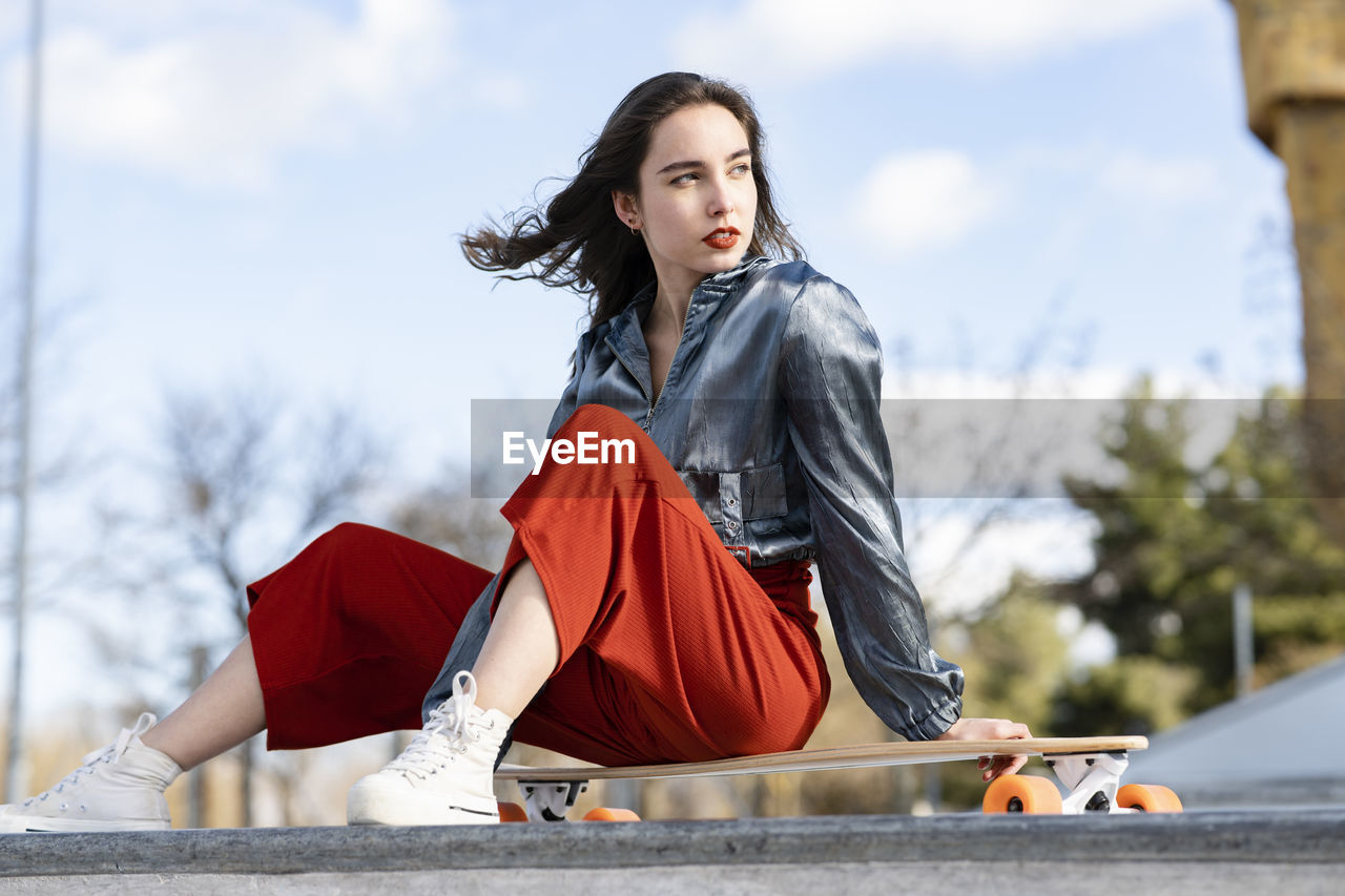 Young attractive woman in stylish clothes sitting on longboard and looking away