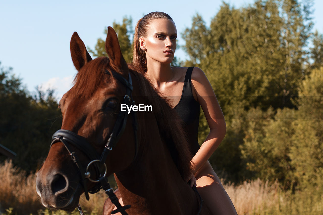 Beautiful young woman riding brown horse on field