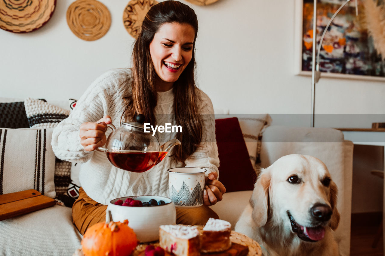 Happy woman pouring coffee in mug while sitting with dog at home