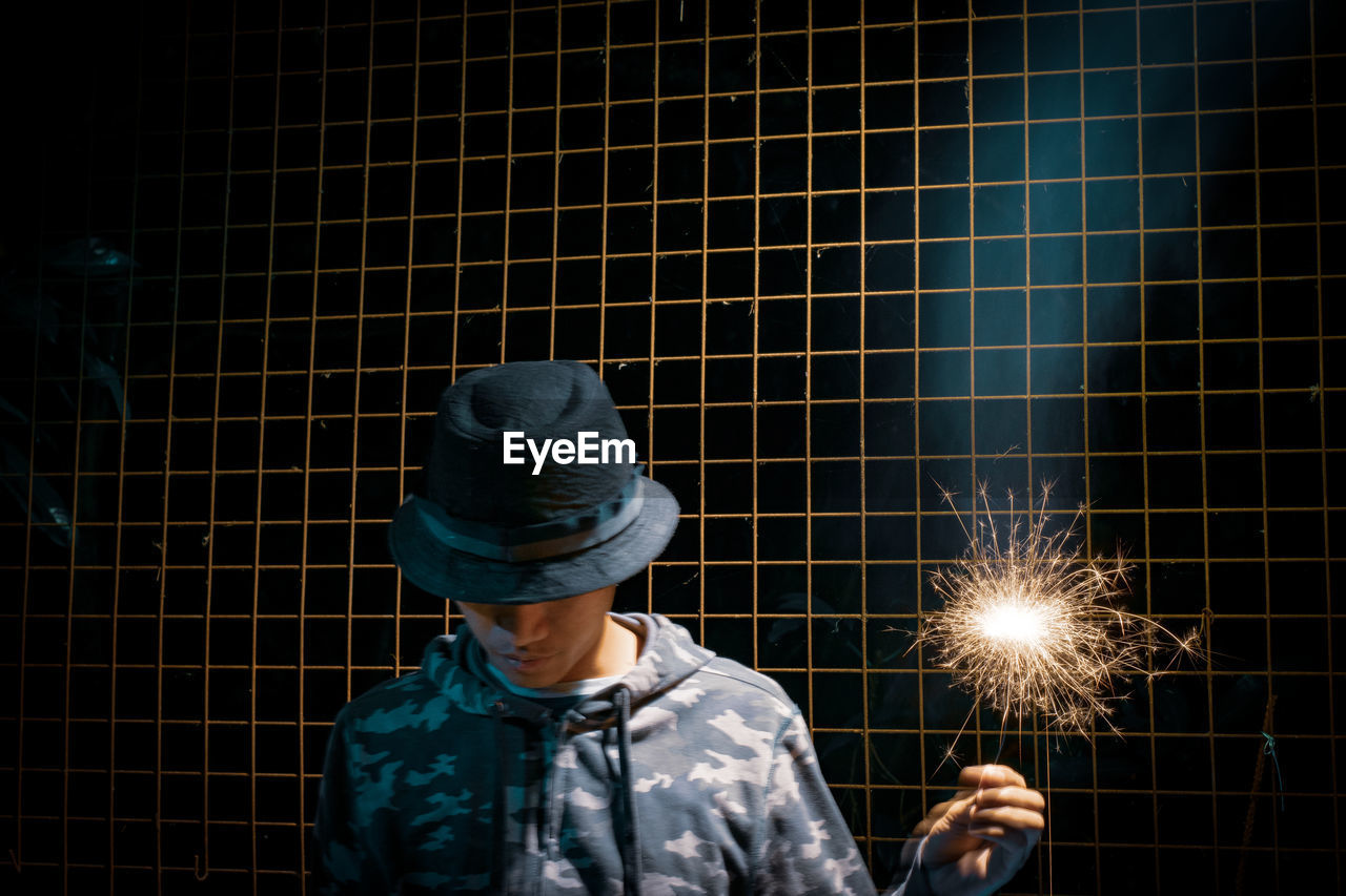 Man wearing hat holding illuminated sparkler against fence