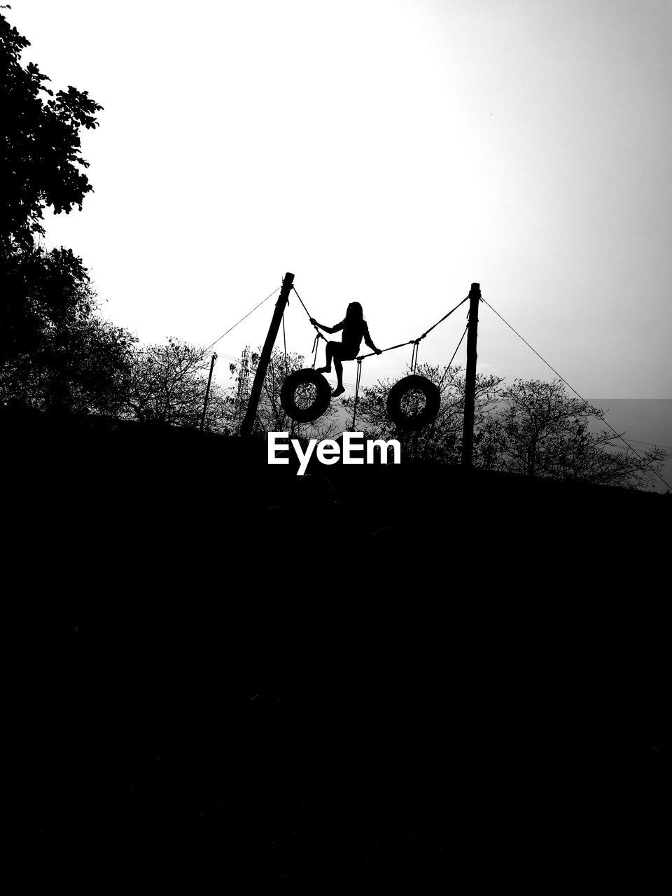 Silhouette woman sitting on rope tied on pole against sky