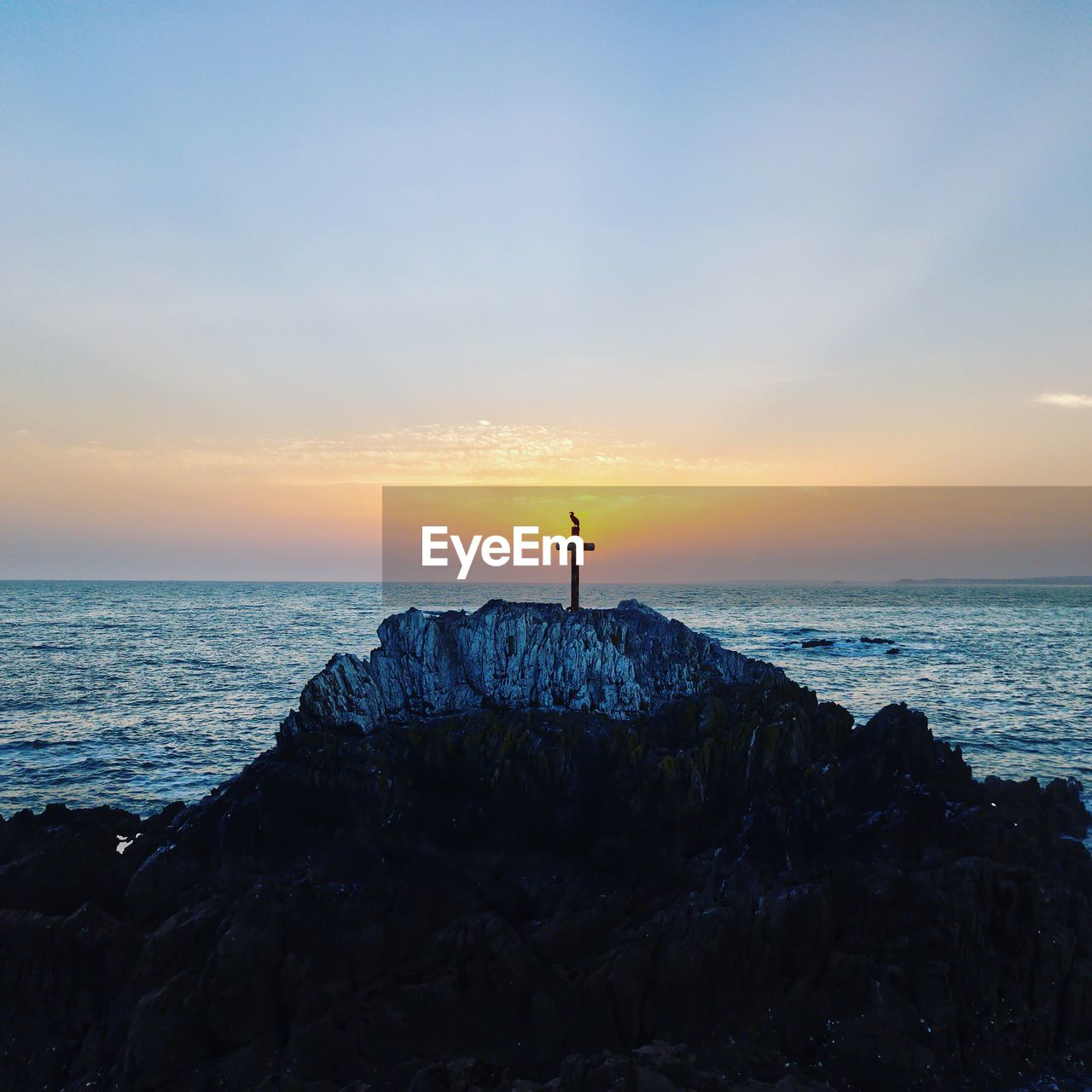 Cross on cliff by sea against sky during sunset