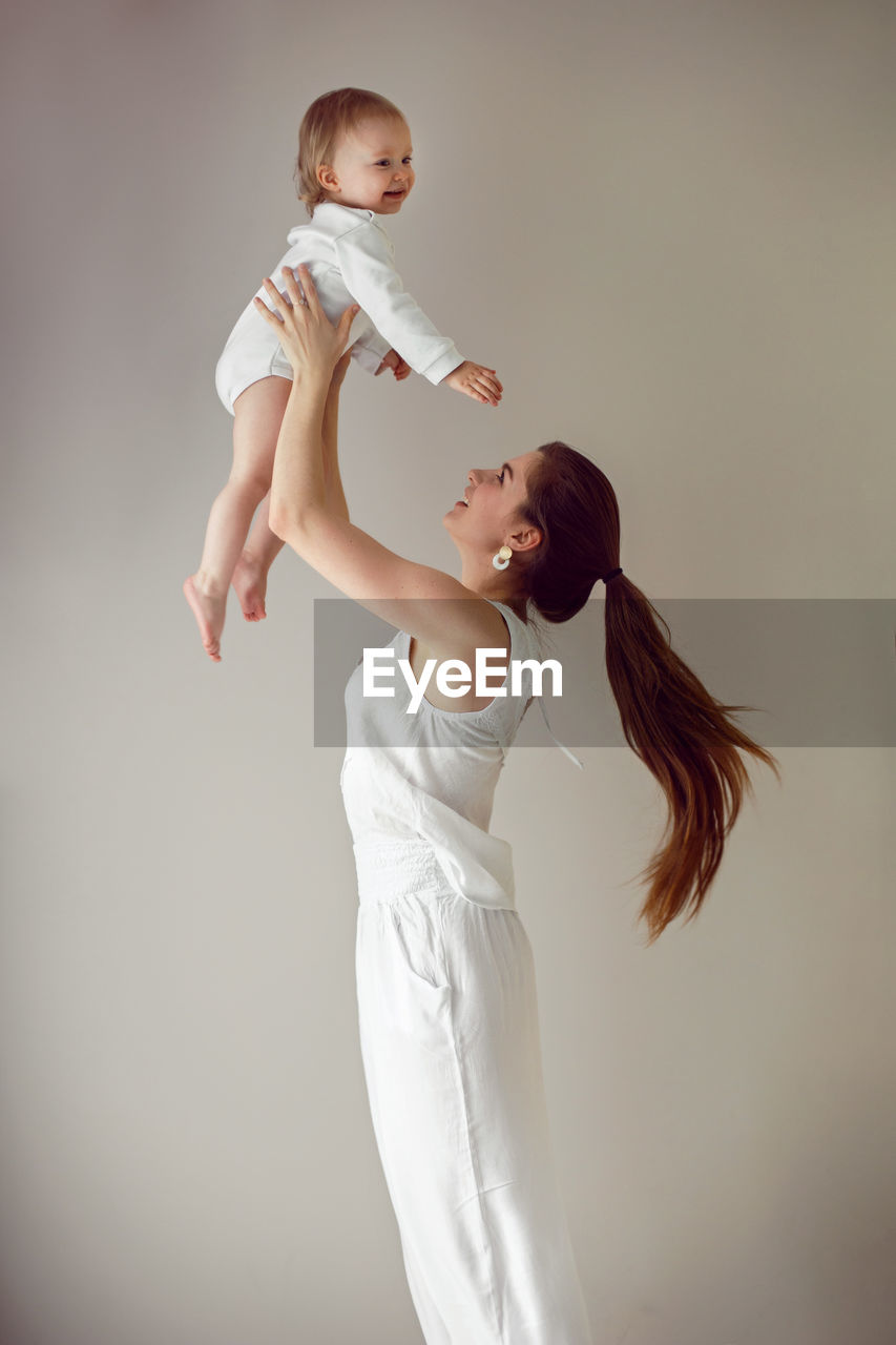 Mom throws her daughter child in her arms stands next to window in white clothes at home in winter