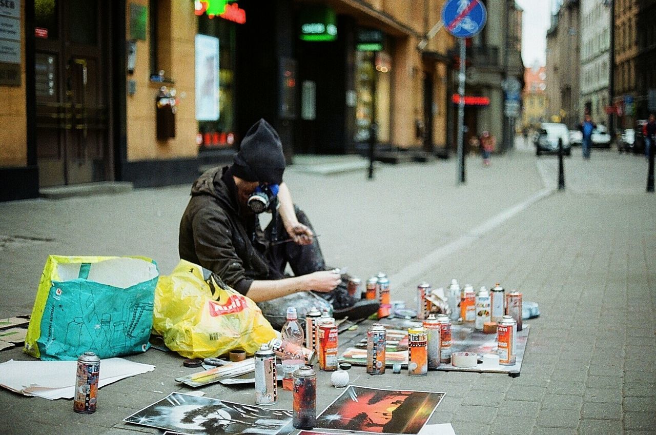 Graffiti artist wearing gas mask sitting with spray paints and paintings on footpath in city