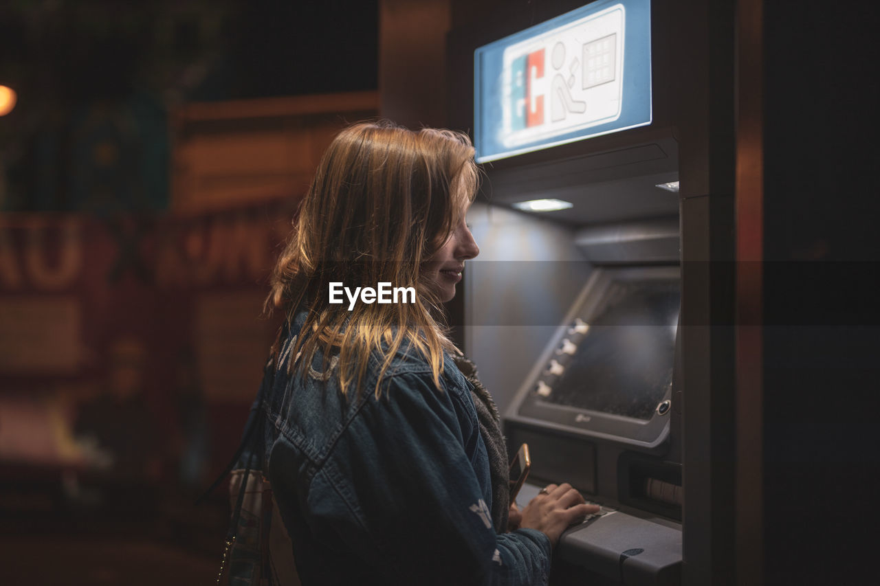 Side view of smiling young woman using atm at night