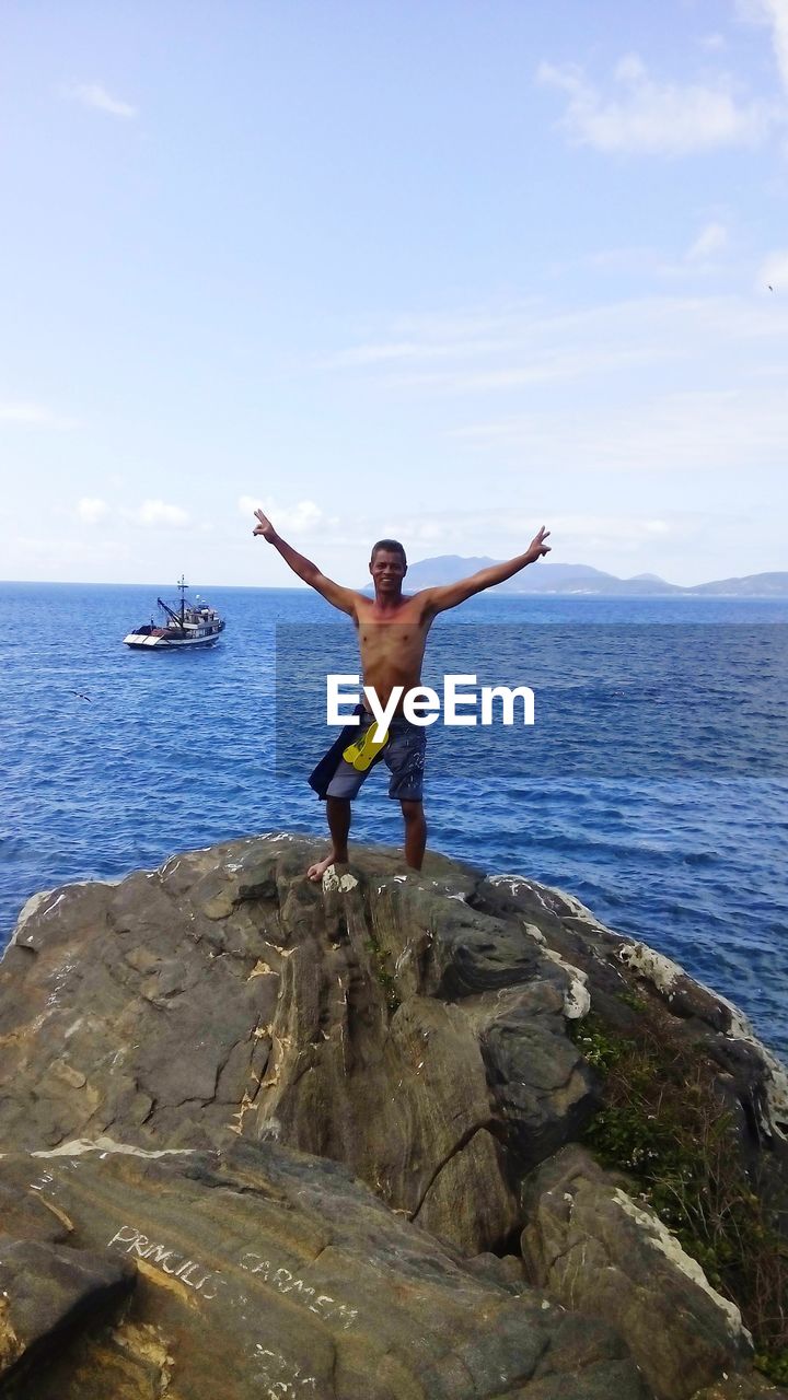 Full length of shirtless man with arms outstretched on rock formation against sea