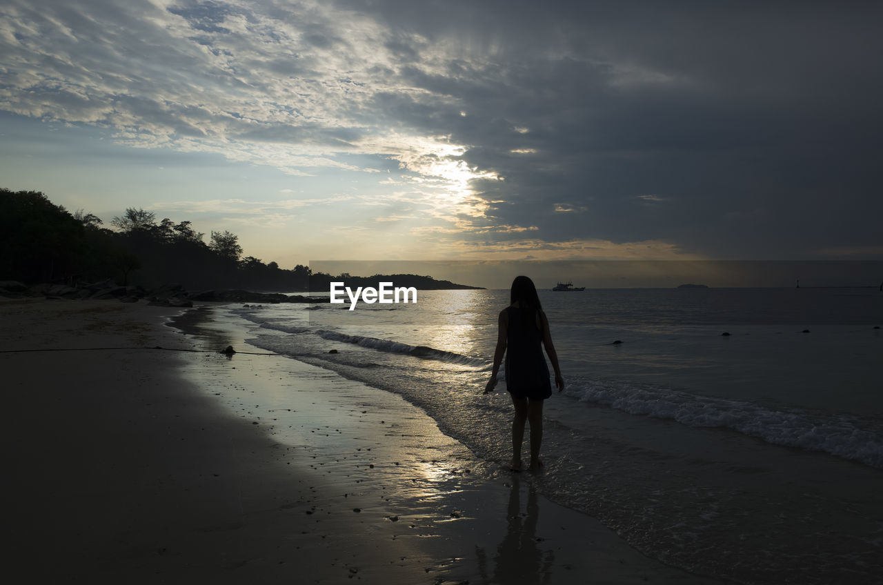 REAR VIEW OF WOMAN WALKING ON BEACH AGAINST SKY DURING SUNSET