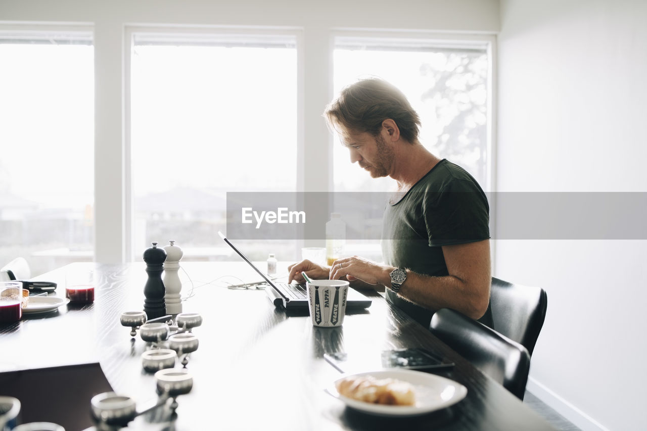 Side view of mature man sitting at dining table using laptop against window