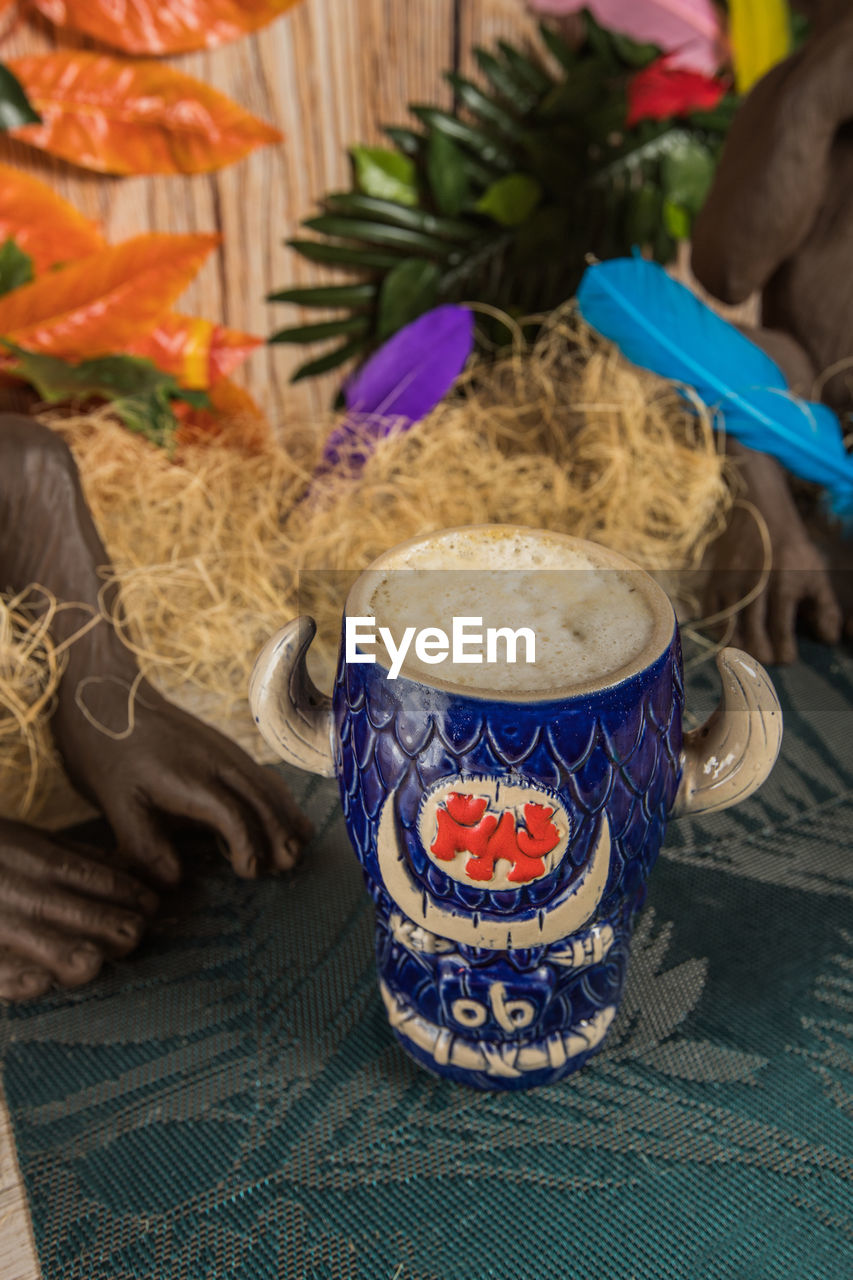 From above of bull shaped tiki mug of alcohol drink with froth placed against dry grass and feathers on blurred background