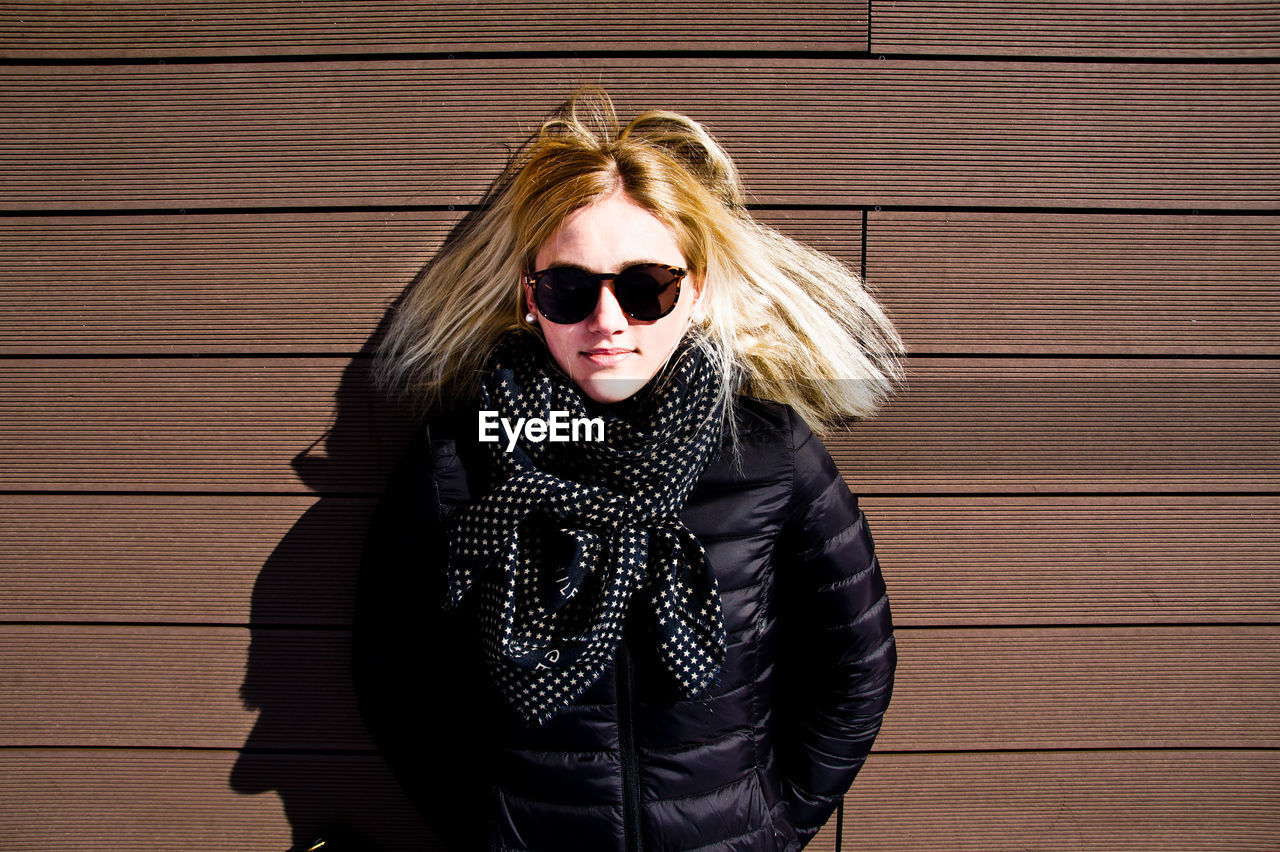 Portrait of young woman wearing sunglasses and warm clothing while standing against wall