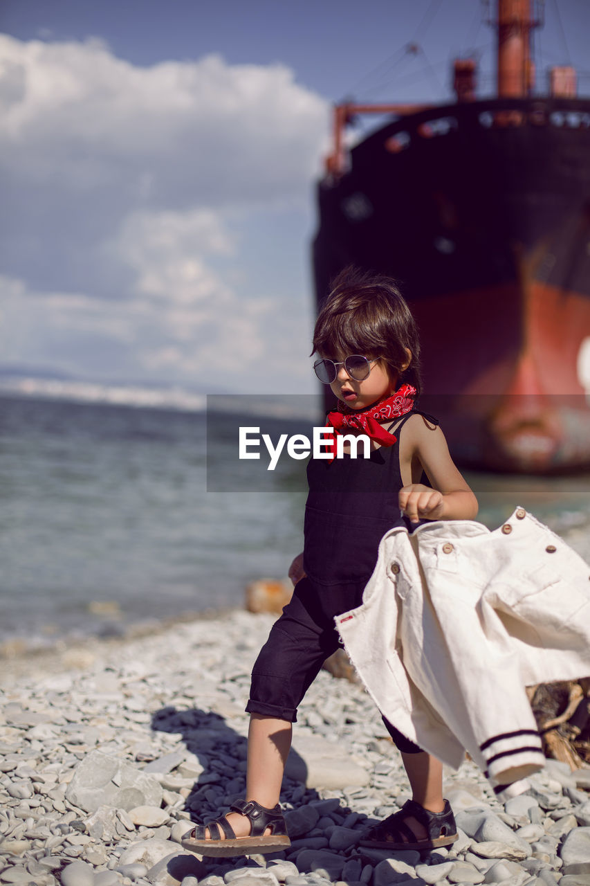 Fashionable boy child with long hair stand on a log next to a large ship that ran aground 