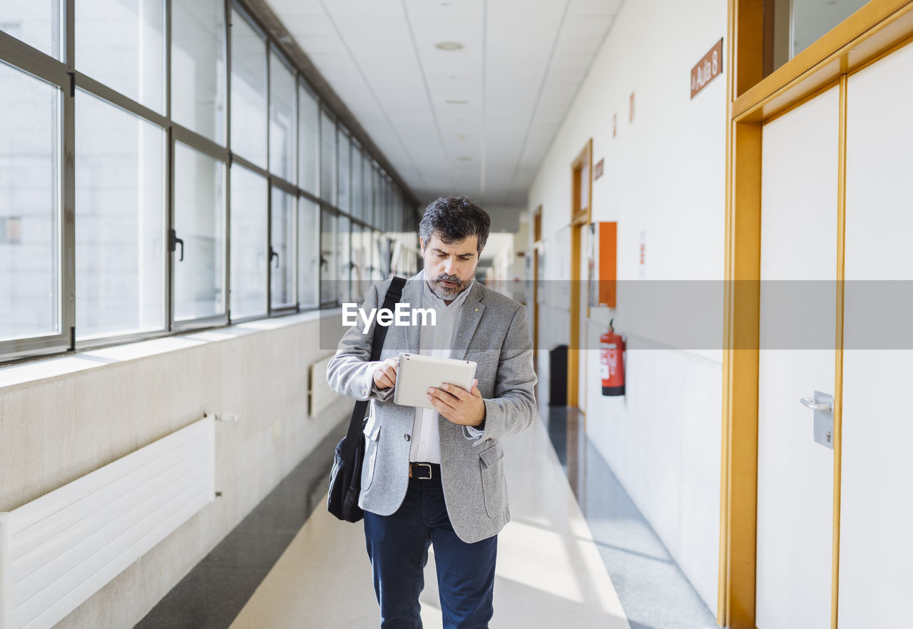 Professor using digital tablet while walking in corridor at university