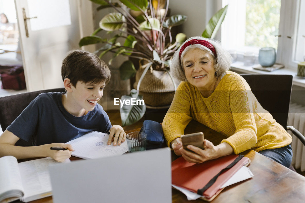 Smiling senior woman using smart phone while sitting with grandson doing homework at home
