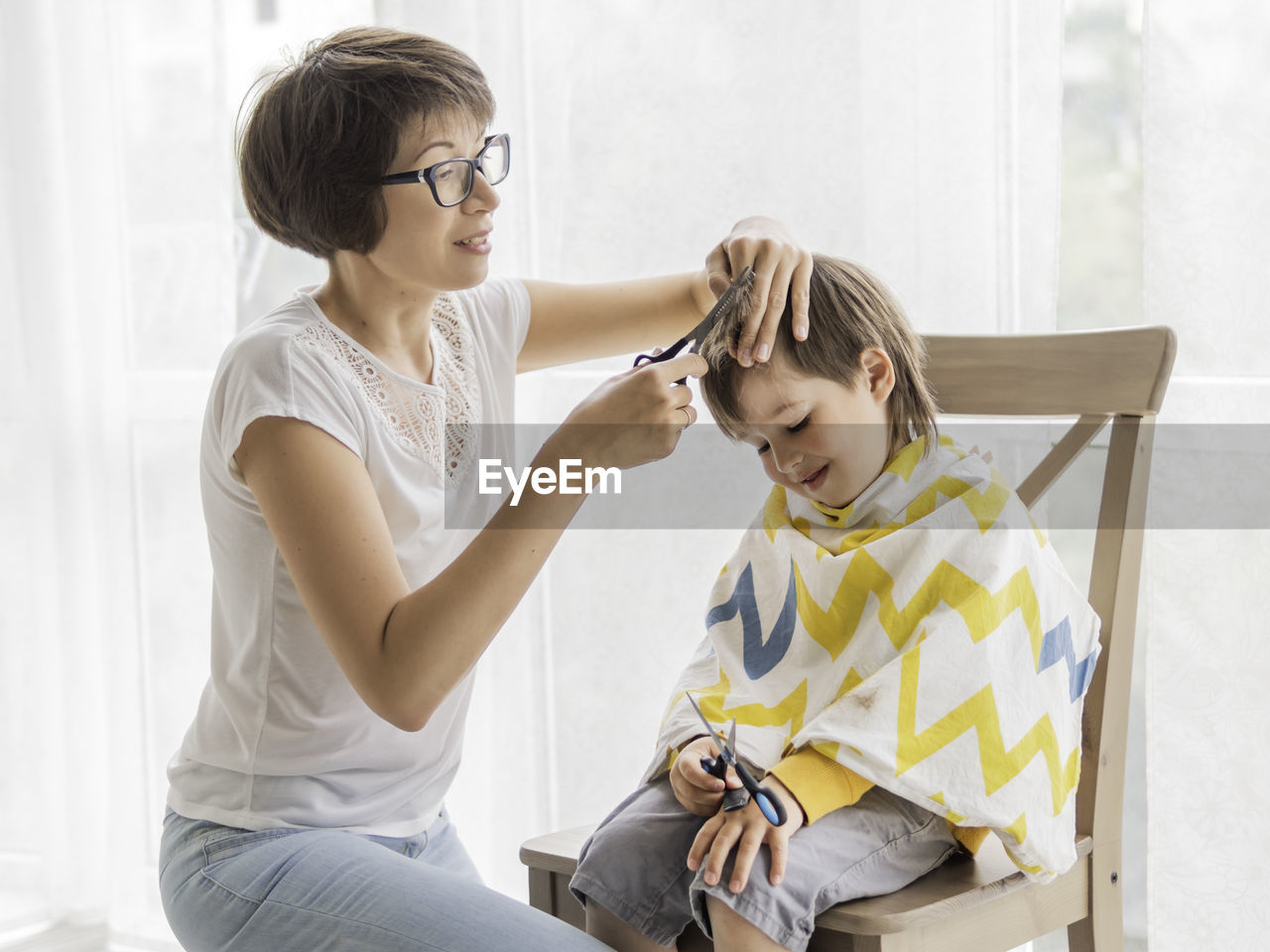 Mother cuts her son's hair by herself. little boy sits, covered with cloth, holds pair of scissors. 