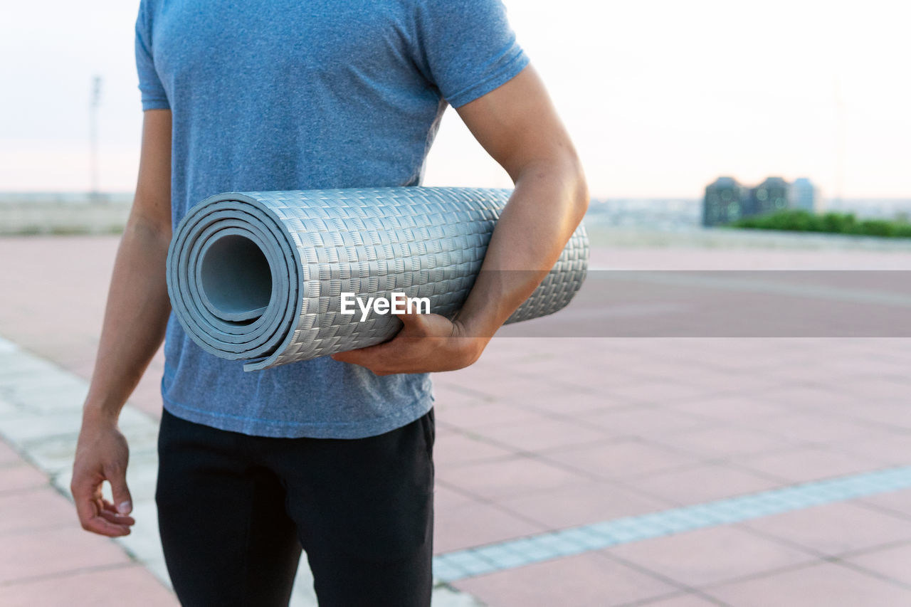 Cropped unrecognizable male in sportswear standing with twisted yoga mat on street and looking away