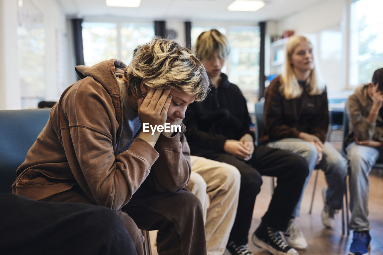 Sad teenage girl with head in hands sitting by male and female friends in group therapy