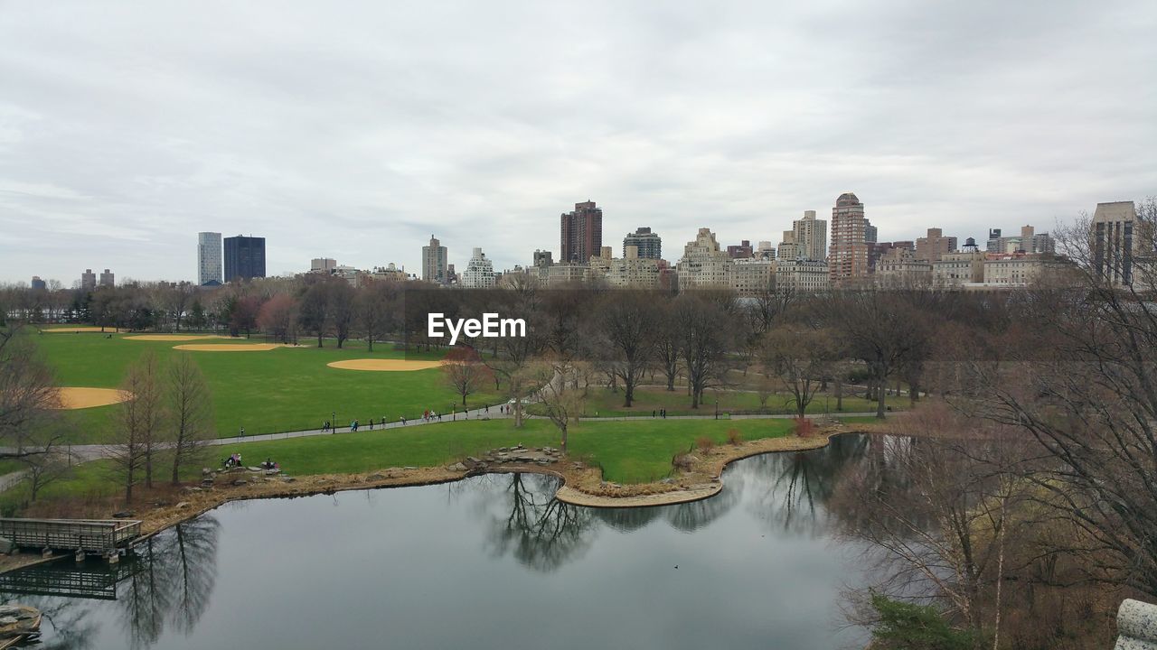 High angle view of park by lake and buildings against cloudy sky