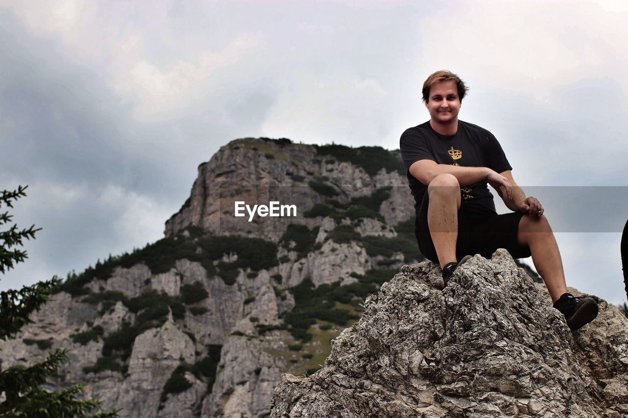Low angle view of smiling man sitting on rock against sky