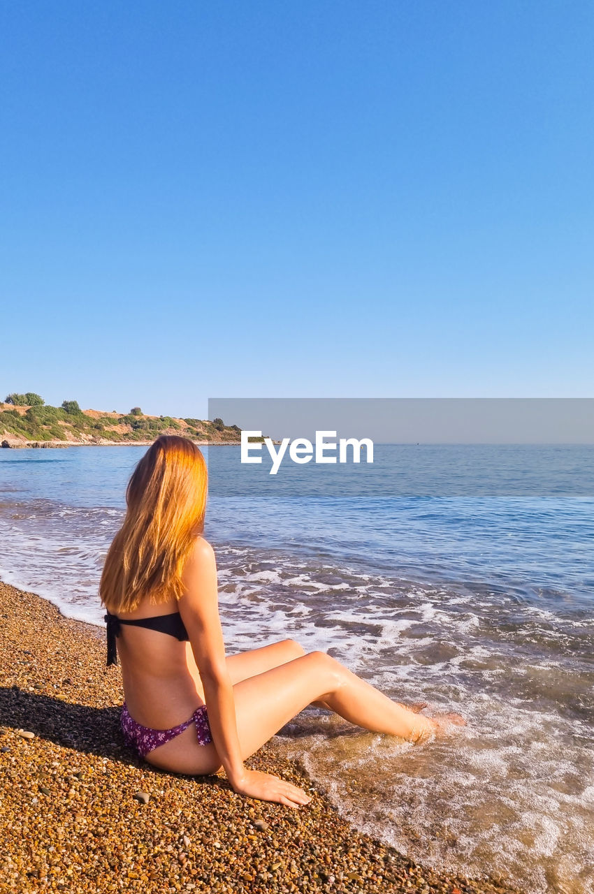 Rear view of woman sitting at beach against clear blue sky
