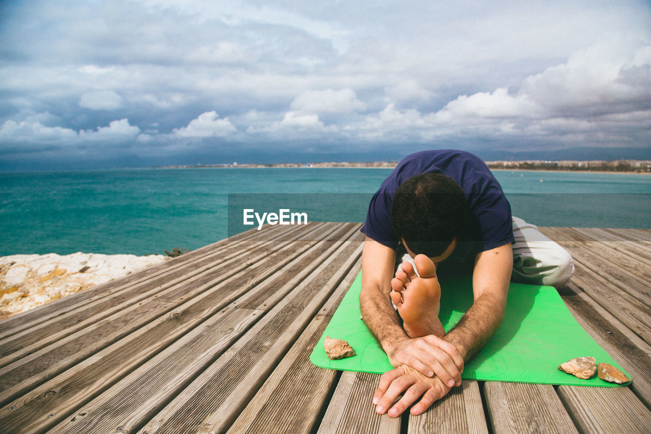 Full length of man performing yoga on jetty over sea against cloudy sky