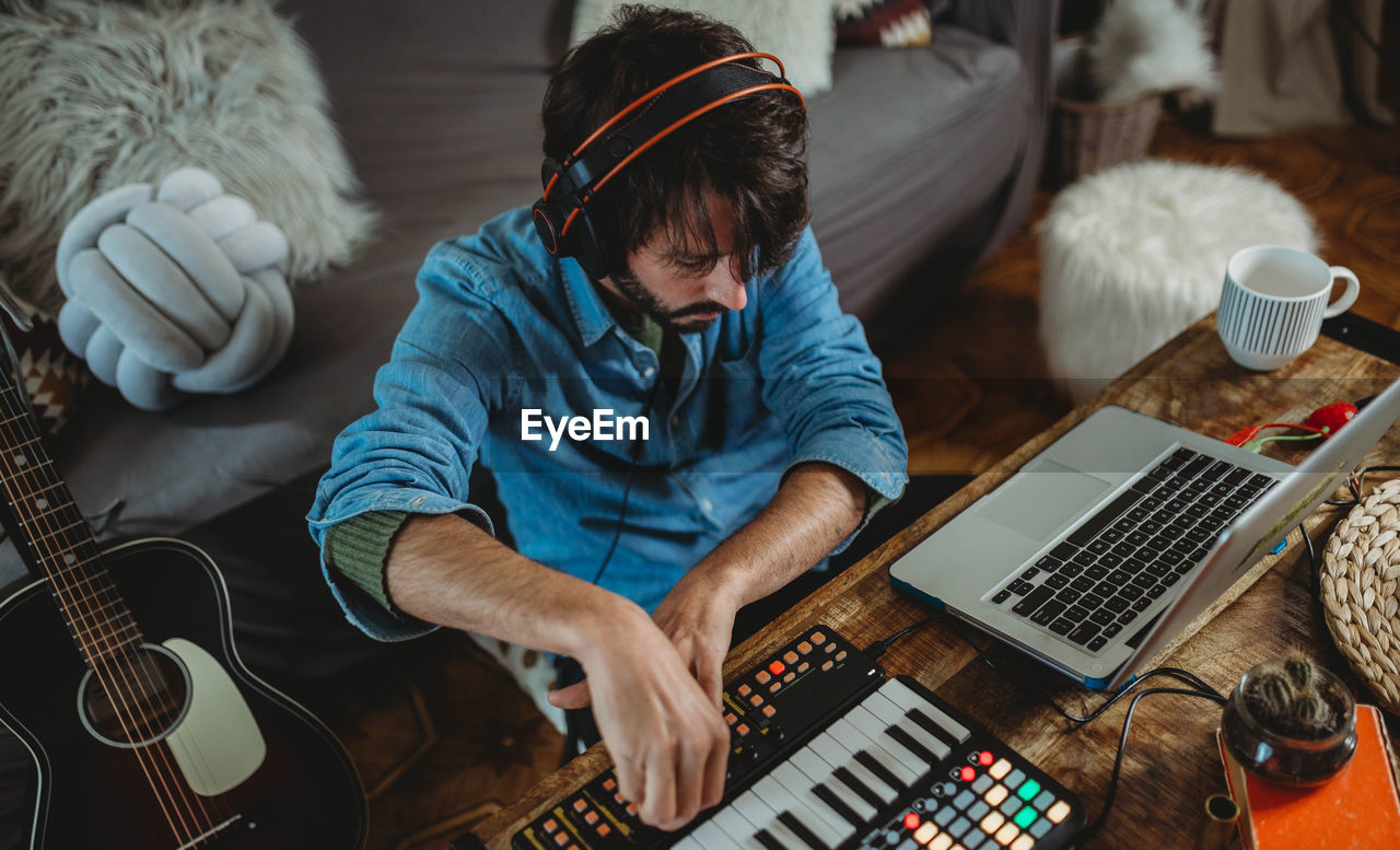 From above happy young man in headphones using synthesizer and laptop at table at home