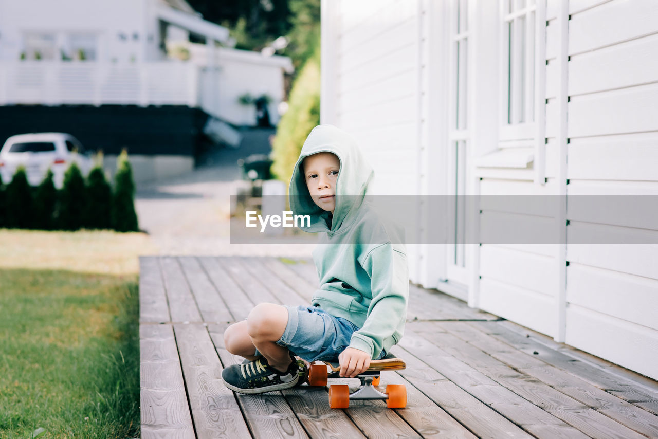 Young boy sat on a skateboard at home in a hoodie