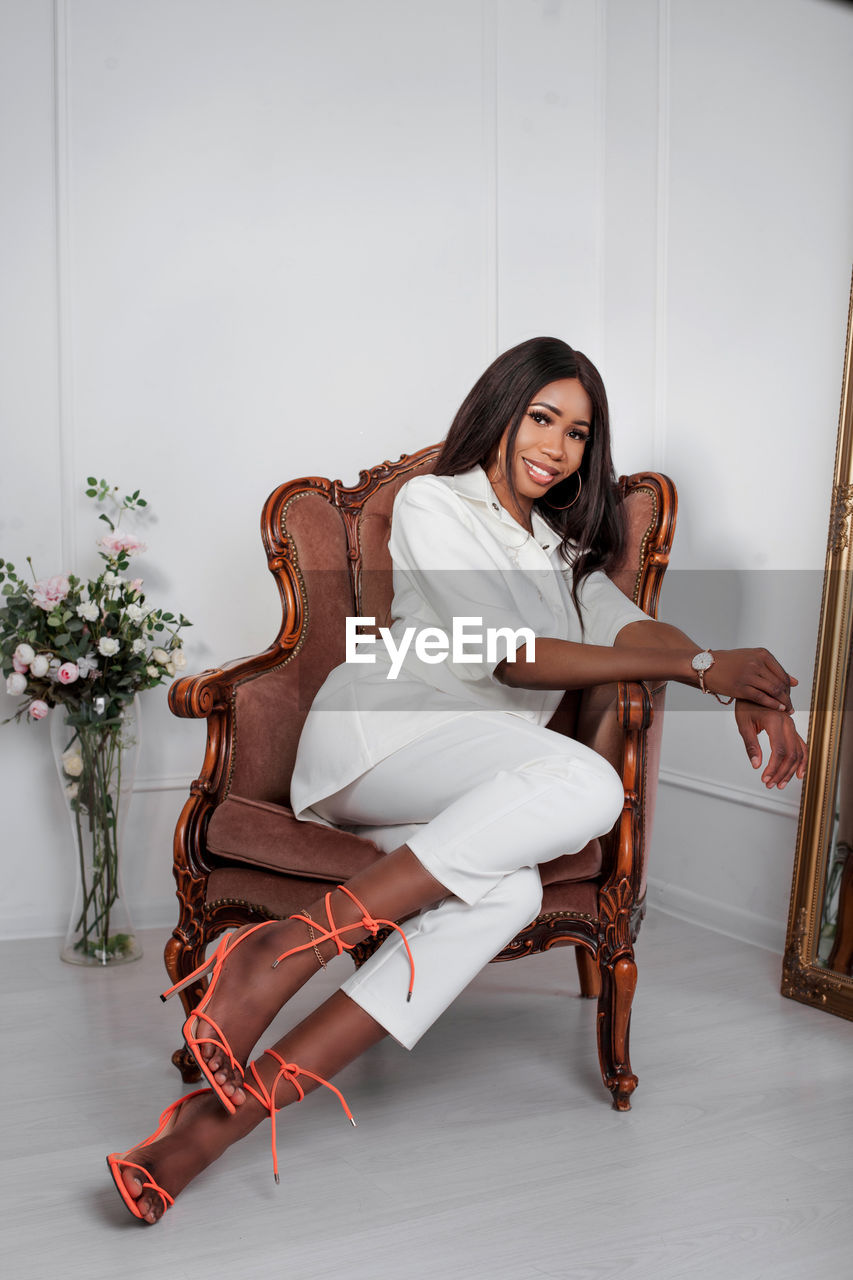 Full length portrait of african nigerian in white pants suit sitting in velvet brown chair in studio