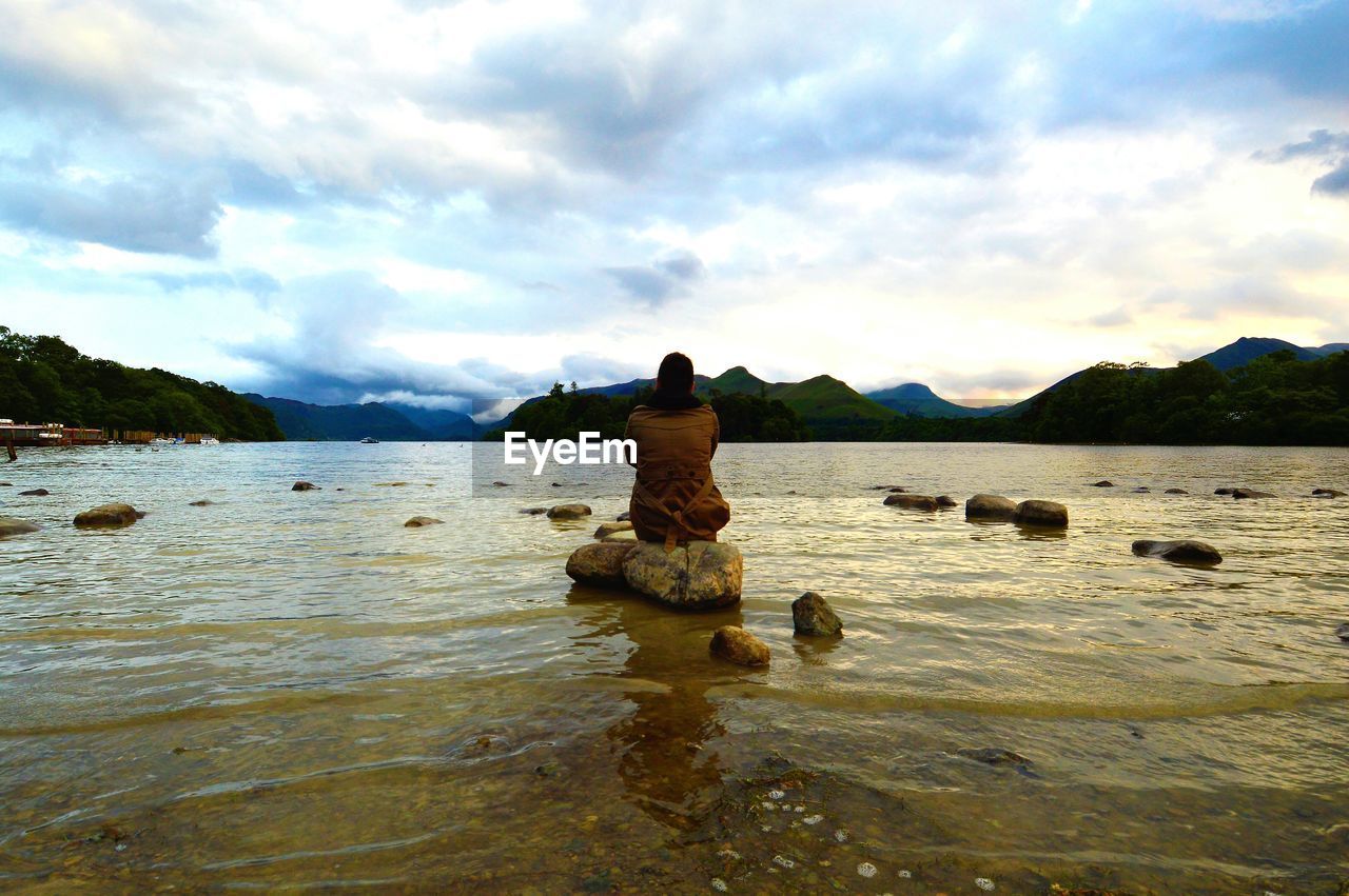 Rear view of man sitting on rock in lake against cloudy sky