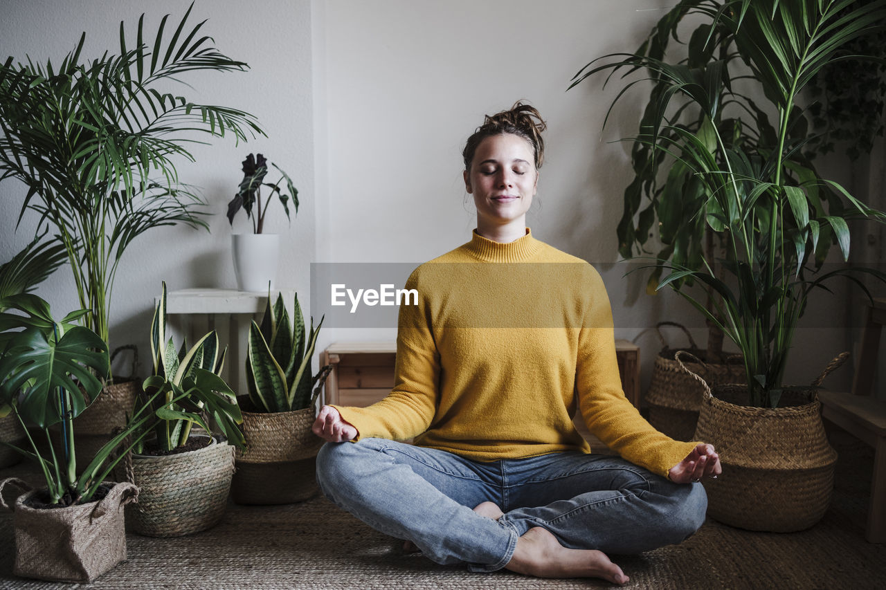 Young woman meditating while sitting on floor by plant at home