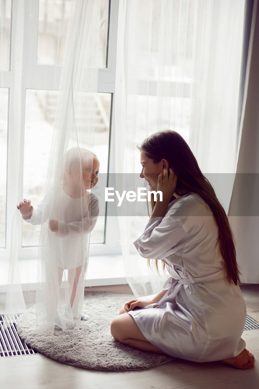 Mother in a white robe sits with a child a blonde daughter at a large window of the house person