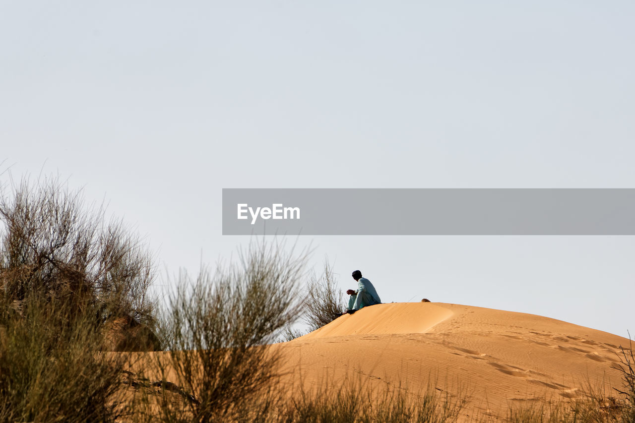 MAN CYCLING ON SAND DUNE AGAINST CLEAR SKY