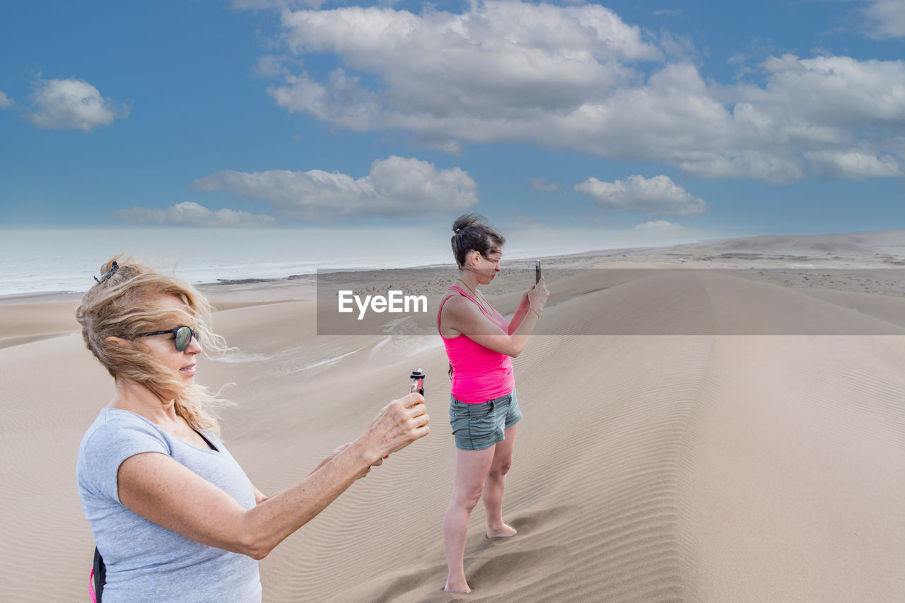 Side view of mature mother and adult daughter taking pictures on top of a big dune with the sea