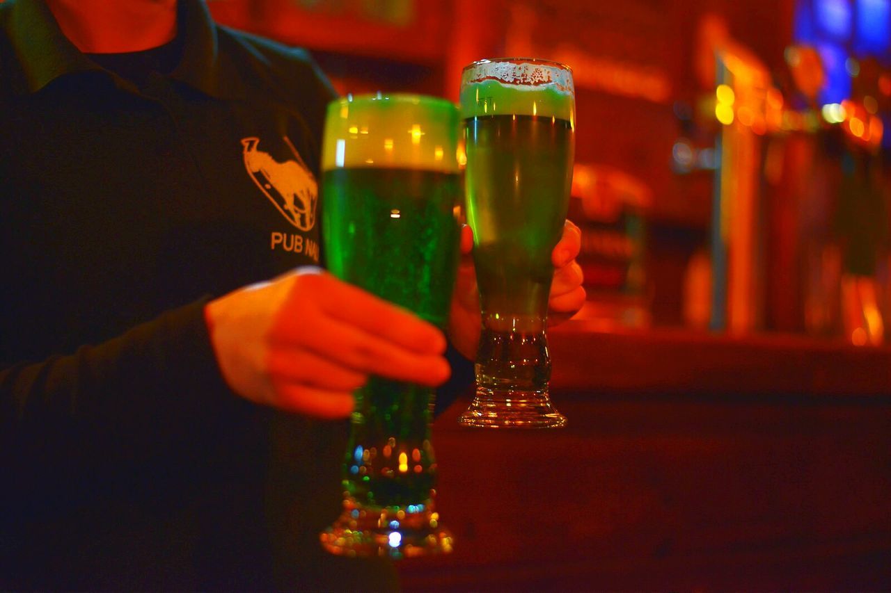 Midsection of bar tender holding green beer glasses in pub