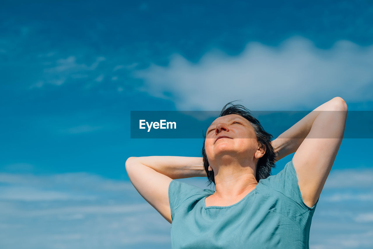 Close-up portrait of an adult smiling woman with her hands behind her head against a blue sky 