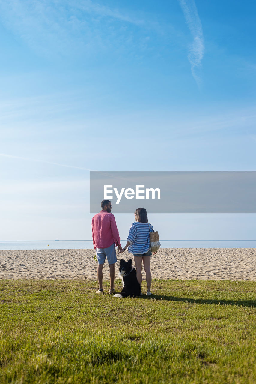 Back view of couple standing and holding hands near their dog on beach