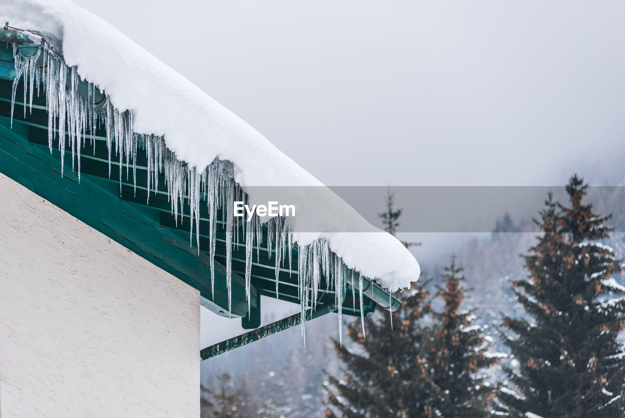 Icicles on snow covered house roof and trees, mountains at the background
